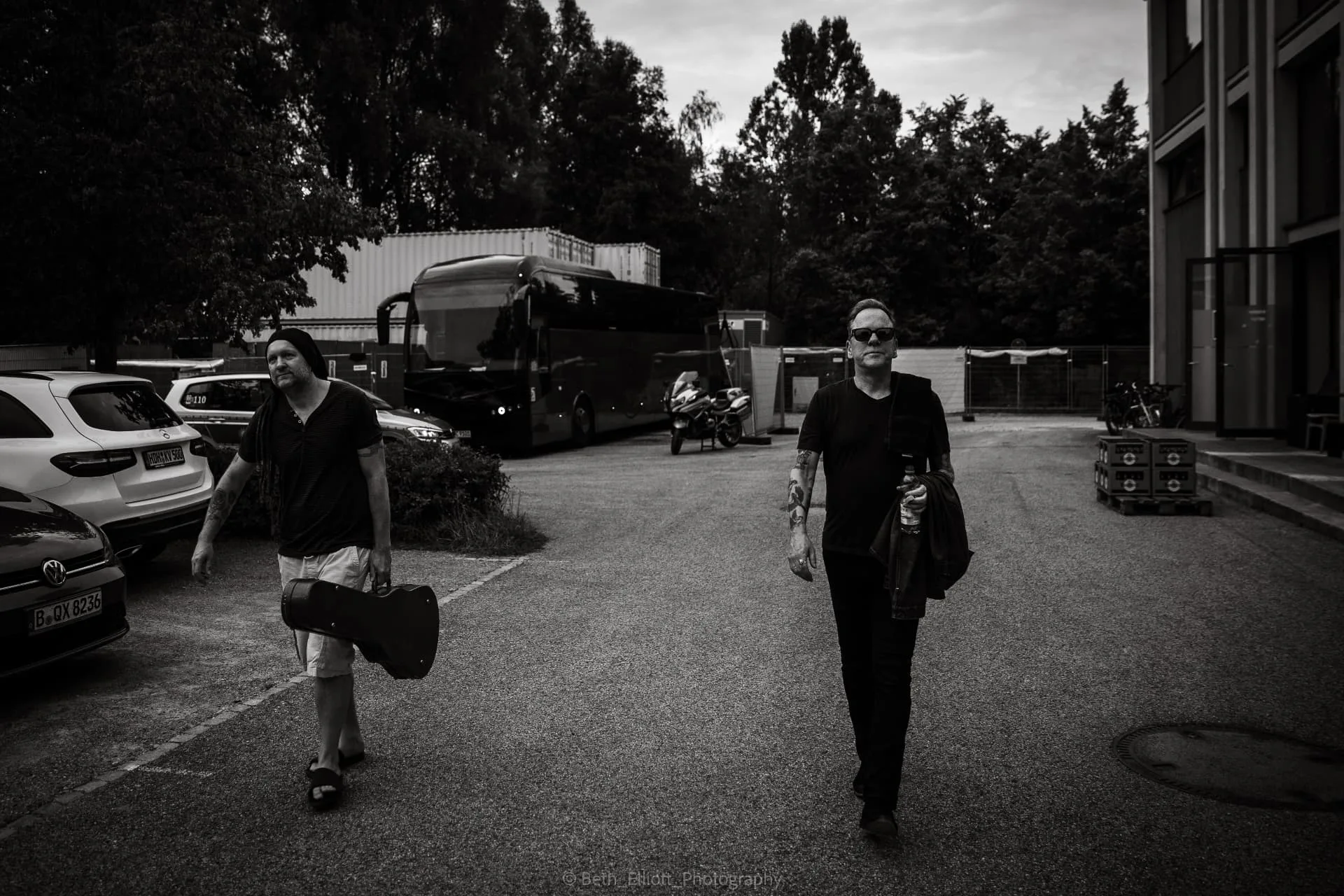 Black-and-white photograph of Nathan Clark and Kiefer Sutherland walking across a backstage car park, with a tour bus behind them